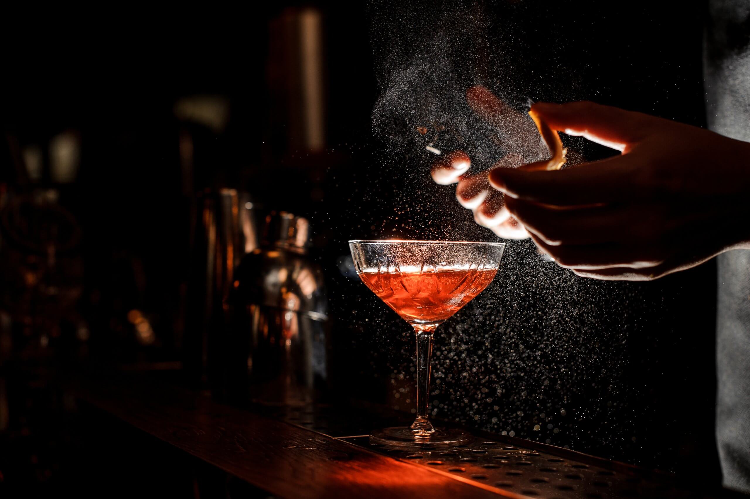 Bartender in black apron and blue shirt sprays an orange peel in cocktail glass with ice at a bar counter