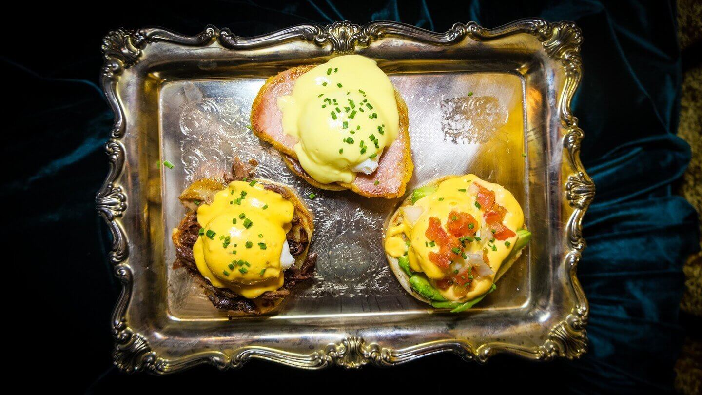 Three eggs benedict with different toppings on a metal tray