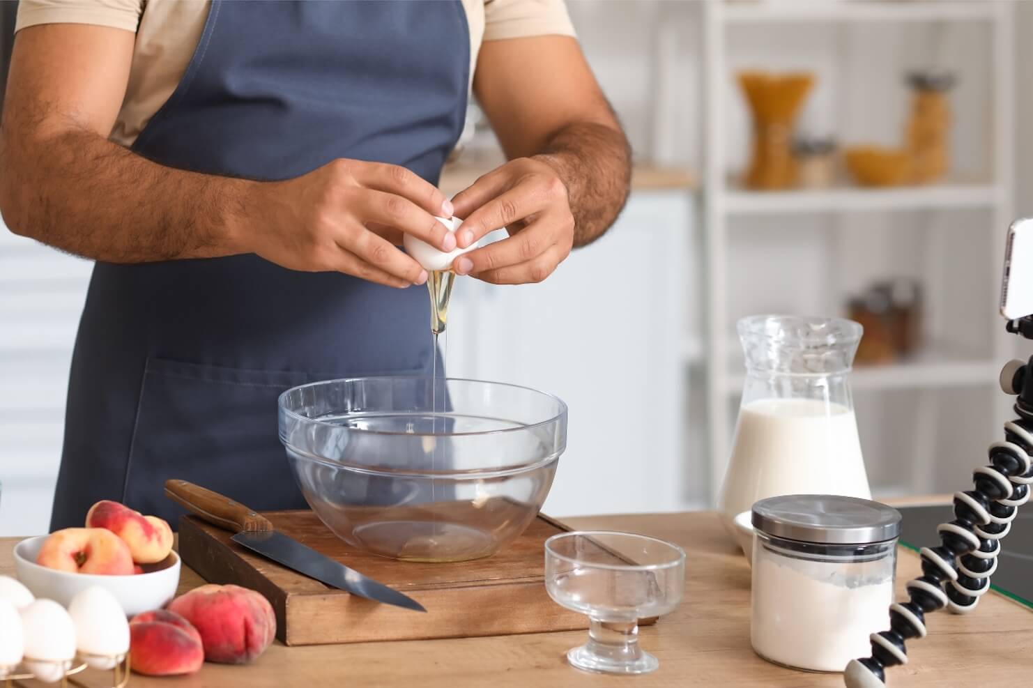 A man in a blue apron breaking an egg into a bowl with a phone in a tripod in front of him