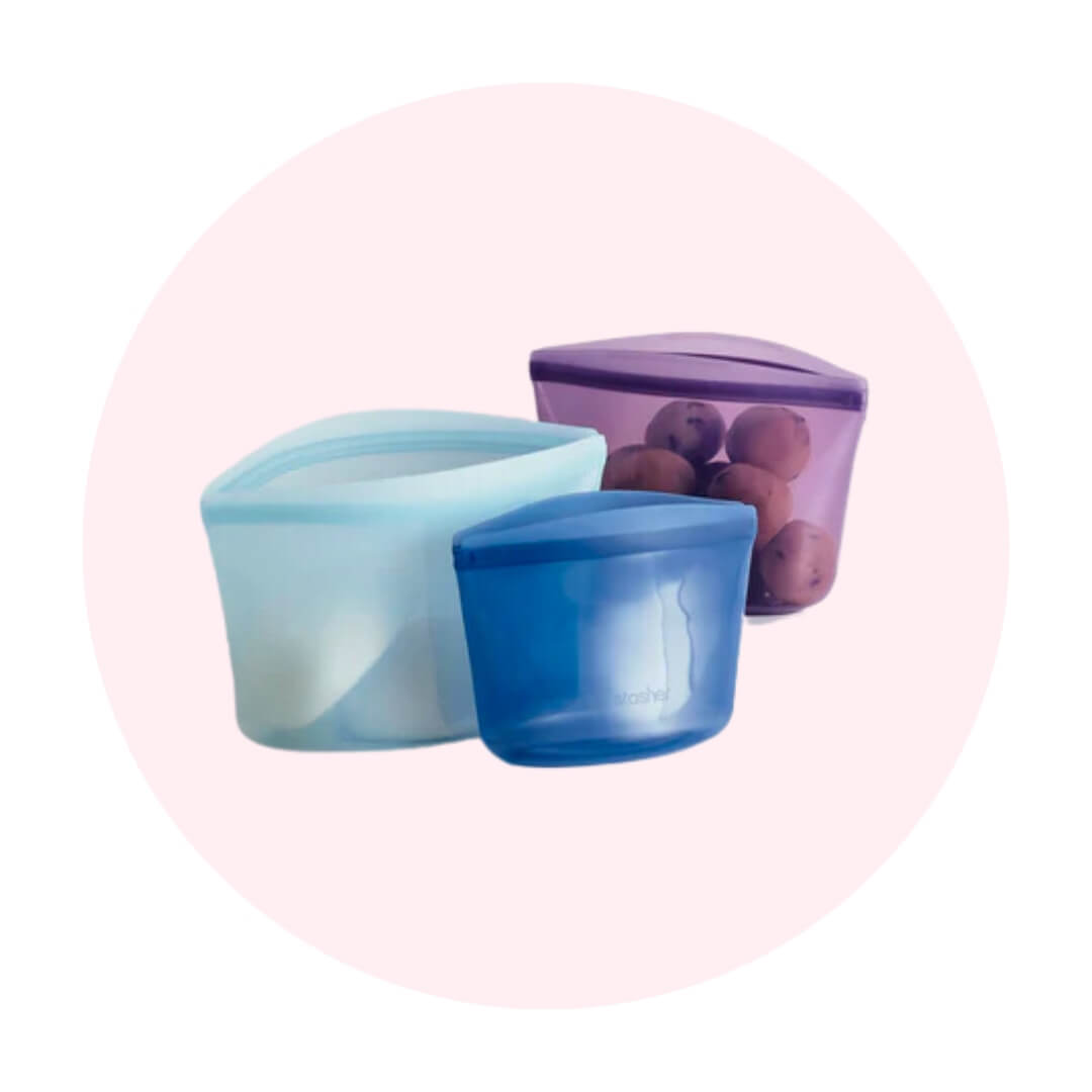 Two blue silicone bags and one purple one with fruit in it on a light pink circle graphic