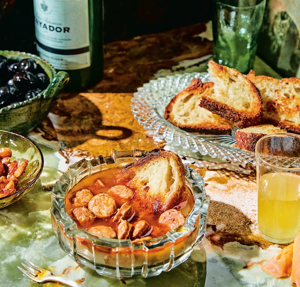 A glass bowl with chorizo and sauce, a plate with toasted bread and green bottles visible in the background
