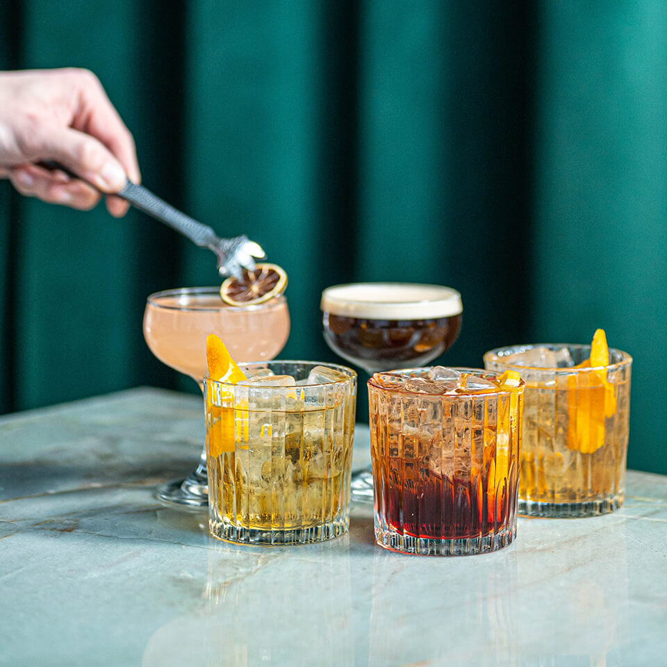 A person's hand using tweezers to garnish a cocktail with a dried citrus wedge, with various other cocktails visible around in on a marble table with a green curtain visible behind it
