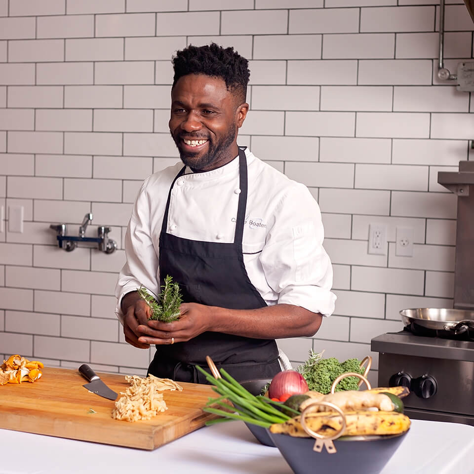 A man, Castro Boateng, laughs while standing in front of a cutting board on a kitchen island in a white-tiled kitchen