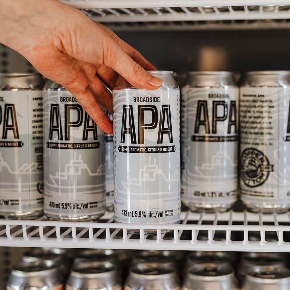 A person's hand reaching for a white can labeled "APA" on a fridge shelf of them