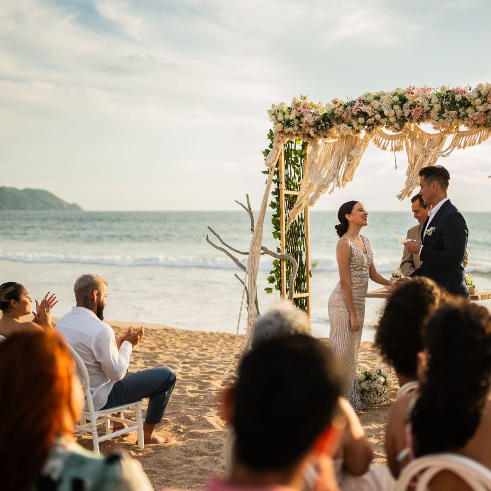 Couple exchanging vows under a floral arch during a beachfront wedding ceremony at sunset.
