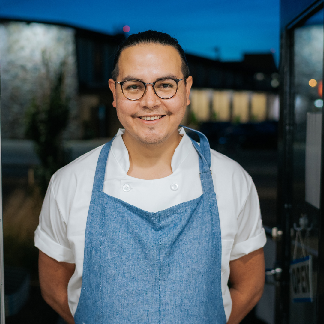 Smiling man wearing glasses and a blue apron standing with his hands behind his back.