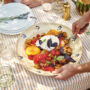 Hands serving a warm tomato and crouton salad topped with burrata and fresh basil on an outdoor table set with plates, glasses and lavender.