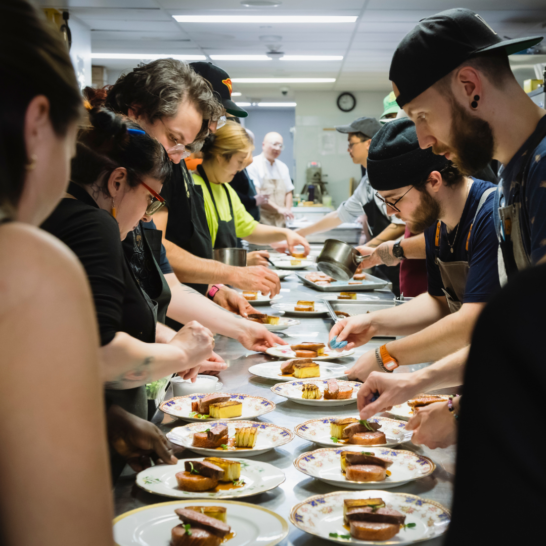 A row of people standing on either side of a counter topped with plates of food. 