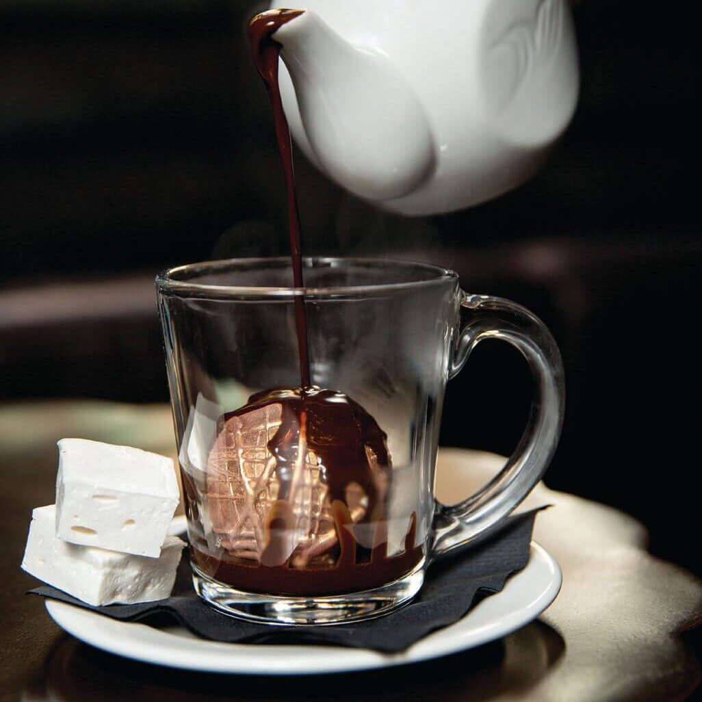 A white teapot pouring chocolate over a hot chocolate bomb with marshmallows on the side.
