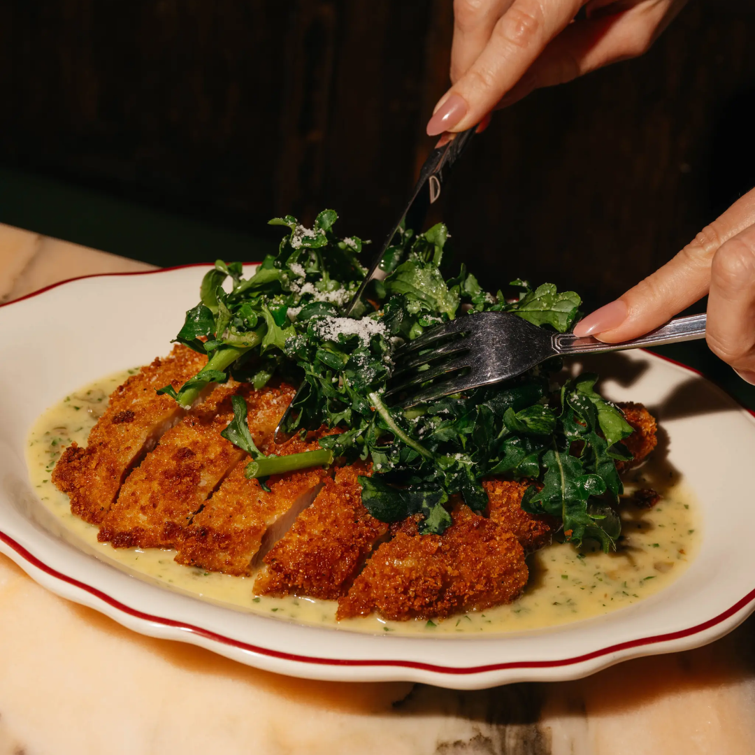 Two hands holding a knife and fork slicing into a fried chicken cutlet.