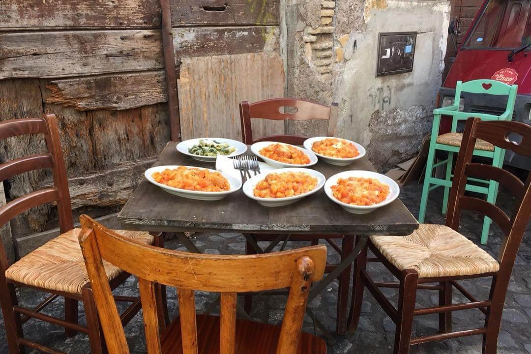 A wooden table laid with different pasta dishes in the centre of mismatched chairs on a cobbled street