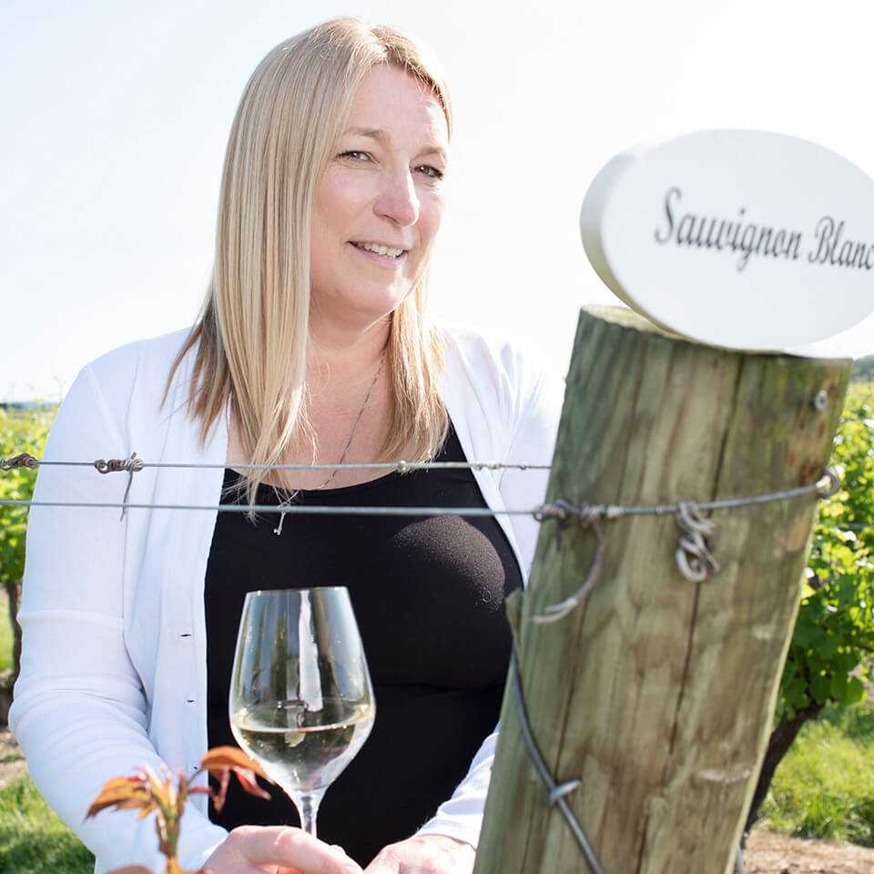 A woman, Andrea Kaiser, stands behind a wooden post with a sign reading "Sauvignon Blanc" in a vineyard while holding a glass of wine