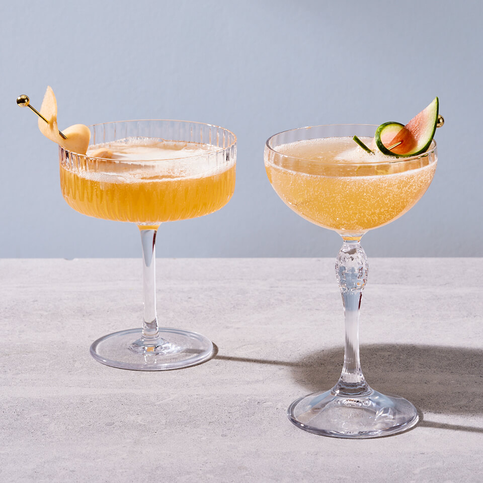 Two light orange cocktails garnished with tropical fruits