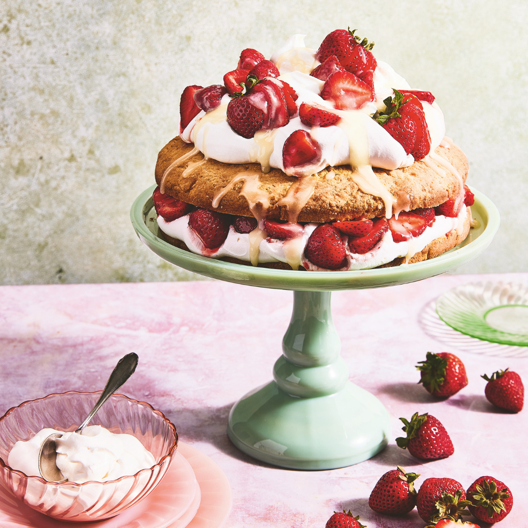 strawberry shortcake layer cake with whipped cream and fresh strawberries ona mint green cake pedestal, surrounded by strawberries and a bowl of whipped cream.