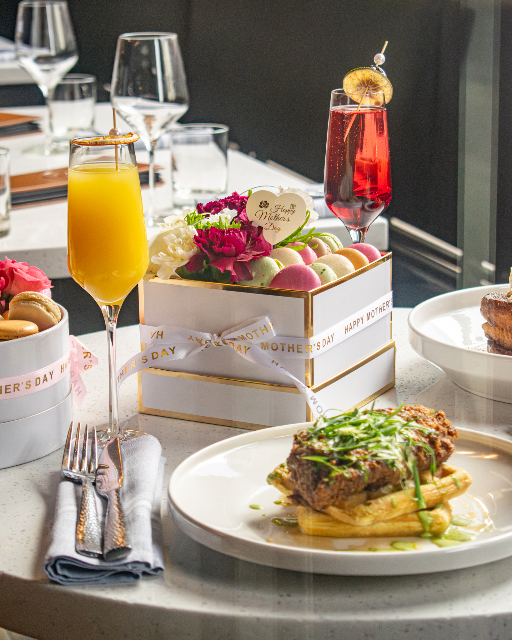 A table with two mimosa glasses, a plate of chicken and waffles. and a box filled with flowers and macarons. 
