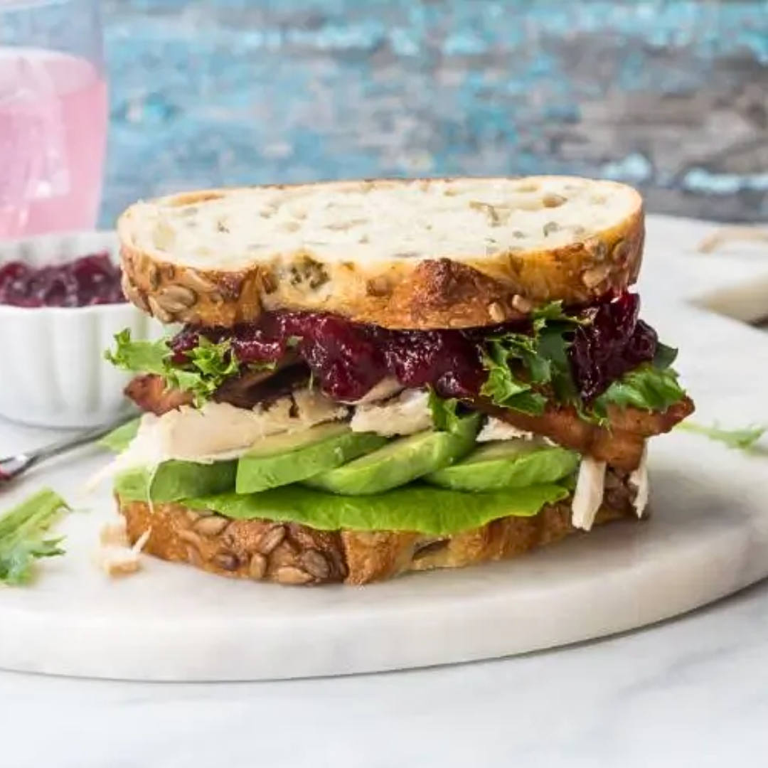 A sandwich filled with cranberry sauce, shredded turkey, lettuce and avocado on a white marble plate. 