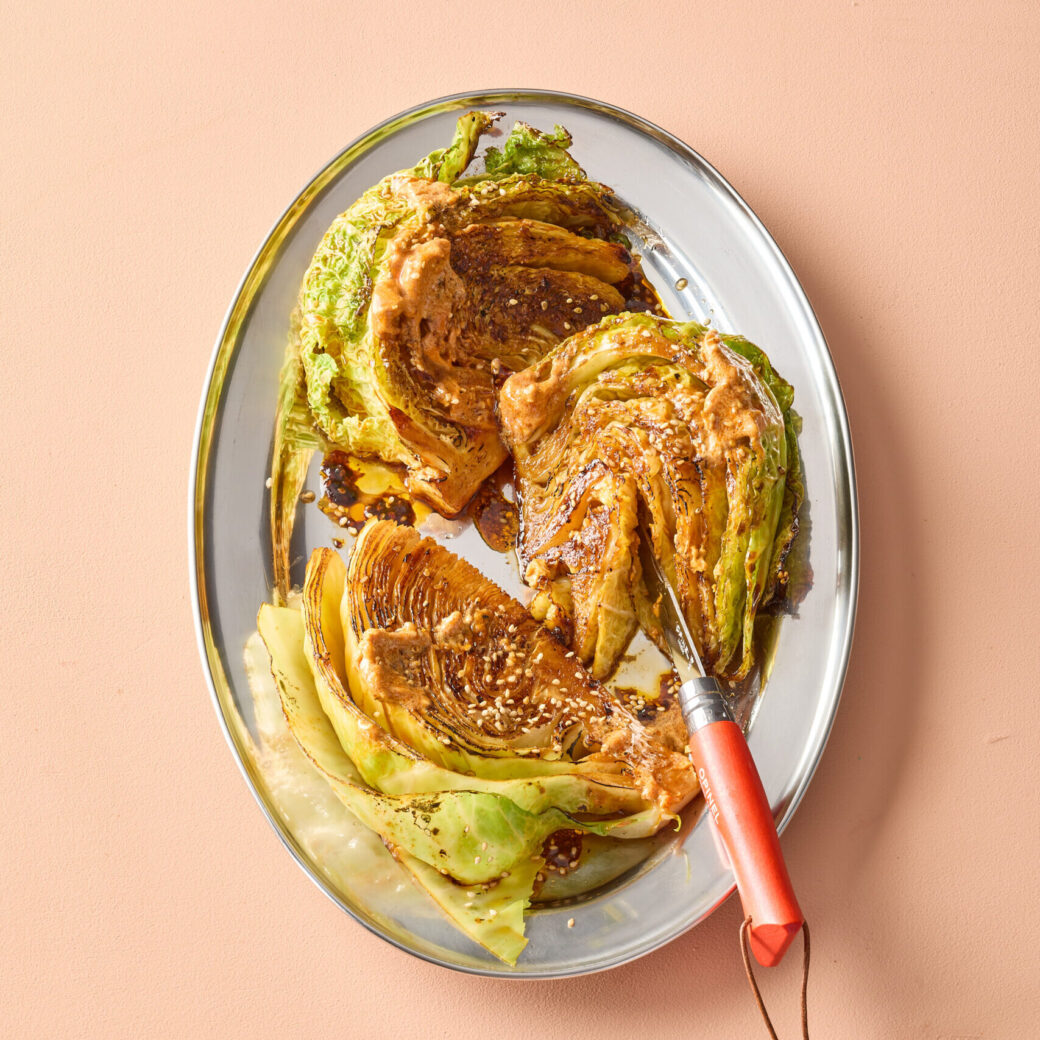 Caramelized cabbage wedges on a platter, drizzled with a creamy sauce and sesame seeds, with a knife resting alongside on a peach-coloured background.