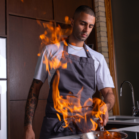 Man in a white shirt and a gray apron holding a pan on fire.