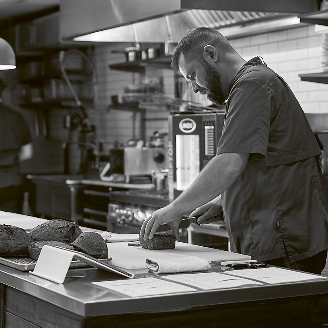 Black and white photo of a man slicing a loaf of bread in the kitchen. 
