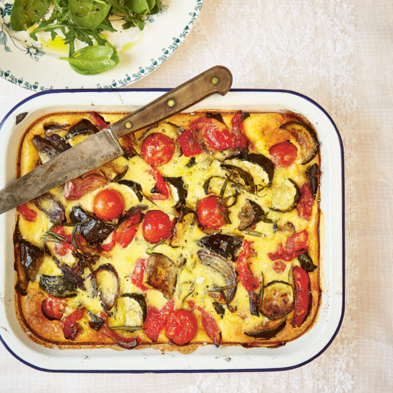 Baked summer vegetable clafoutis in a white dish, studded with roasted eggplant, zucchini, peppers and cherry tomatoes, with a knife resting on top and a simple green salad and glass of white wine alongside.