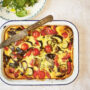 Baked summer vegetable clafoutis in a white dish, studded with roasted eggplant, zucchini, peppers and cherry tomatoes, with a knife resting on top and a simple green salad and glass of white wine alongside.