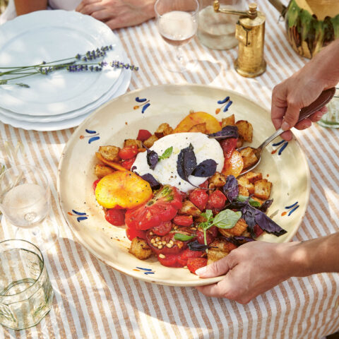Hands serving a warm tomato and crouton salad topped with burrata and fresh basil on an outdoor table set with plates, glasses and lavender.