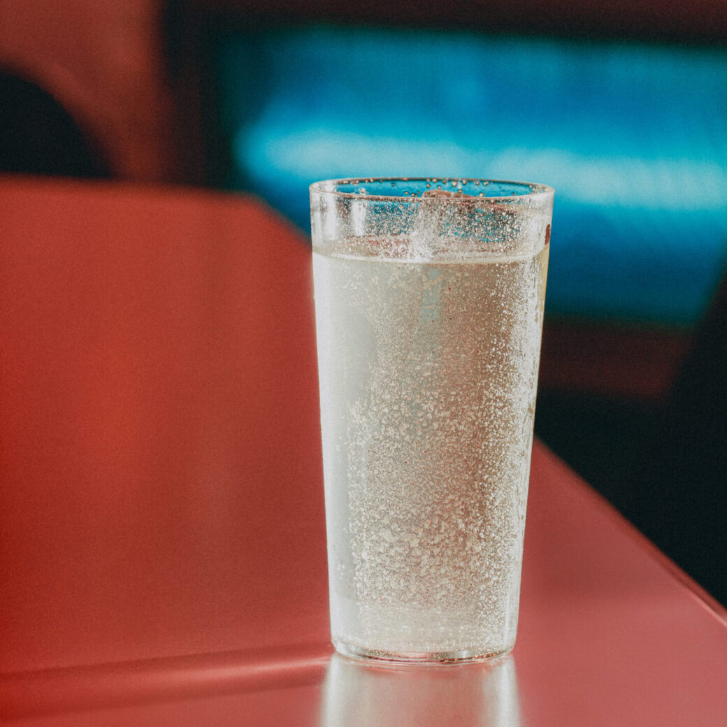 A clear sparkling cocktail in a tall glass sits on a glossy red table with warm moody lighting and a softly glowing blue background.